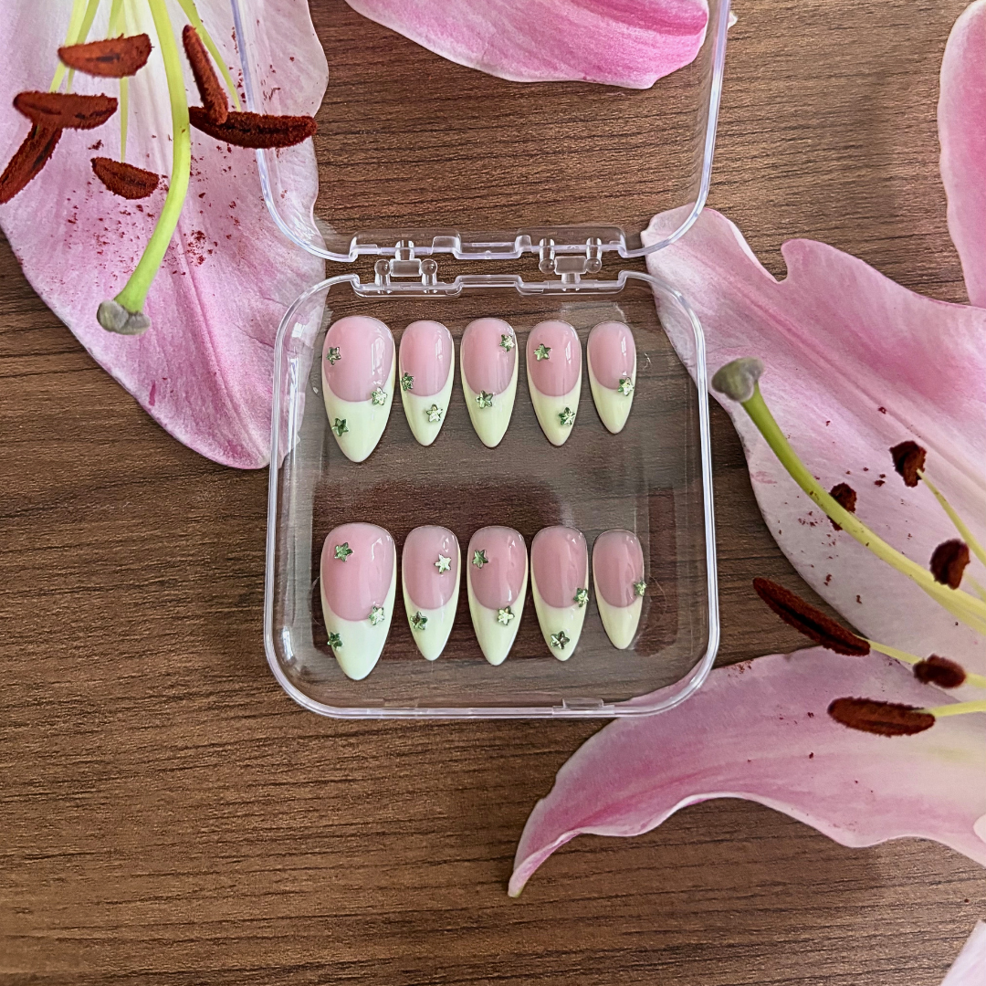 Set of pink and green nail art designs in a clear case on a wooden surface with pink lily flowers.